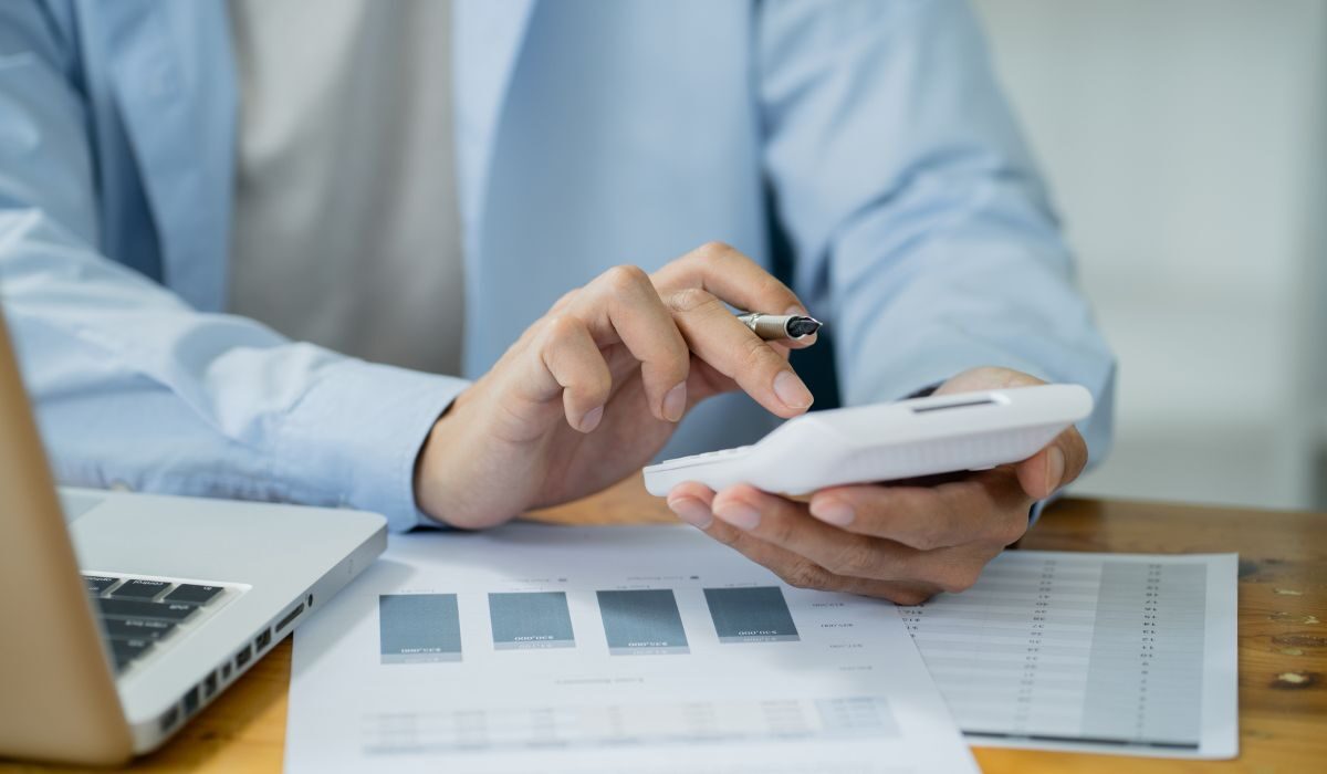 man calculating an invoice through calculator with a laptop on table and some papers